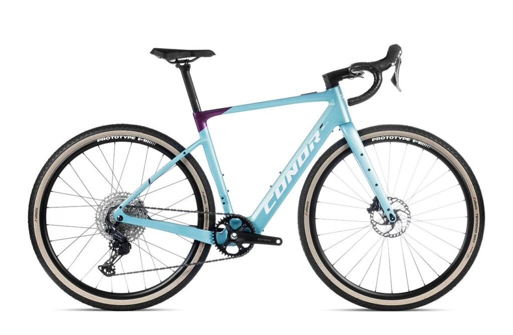 Vista lateral completa de la bicicleta eléctrica de gravel Conor E‑SELVA con cuadro de carbono en color turquesa y púrpura, montada con grupo Shimano GRX y ruedas Prototype de perfil bajo