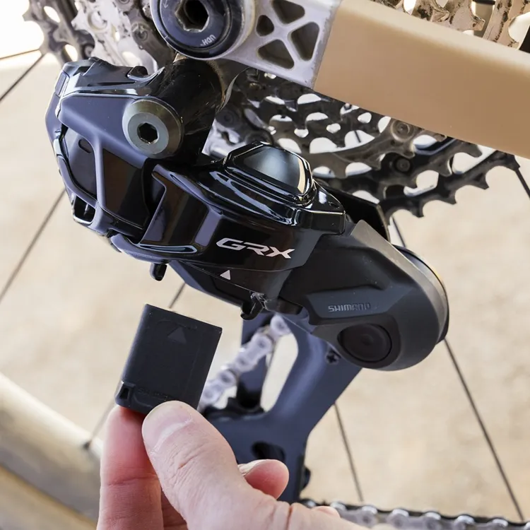 Primer plano del nuevo desviador trasero Shimano GRX Di2 827 de 12 velocidades montado en una bicicleta de gravel, mientras una mano sostiene una batería extraíble para su instalación o carga. El componente muestra el logo GRX y detalles del mecanismo electrónico.