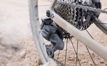 Imagen de un desviador trasero Shimano GRX Di2 de 12 velocidades cubierto parcialmente de polvo, montado en una bicicleta de gravel. Se aprecia el cassette de amplio rango y el disco de freno trasero, con fondo de terreno rocoso que sugiere condiciones off-road exigentes.