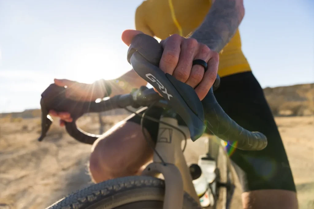 Primer plano de un ciclista sujetando la maneta de cambio Shimano GRX Di2 mientras pedalea por un terreno árido. La maneta, con el logo GRX visible, está montada en un manillar de gravel, iluminada por la luz del sol al atardecer.
