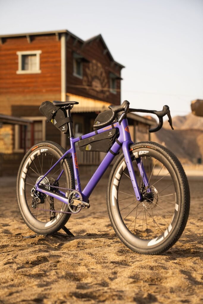 Versión Malva de la Megamo West 2026 equipada para bikepacking, con bolsas delanteras y traseras, en un entorno tipo desierto.
