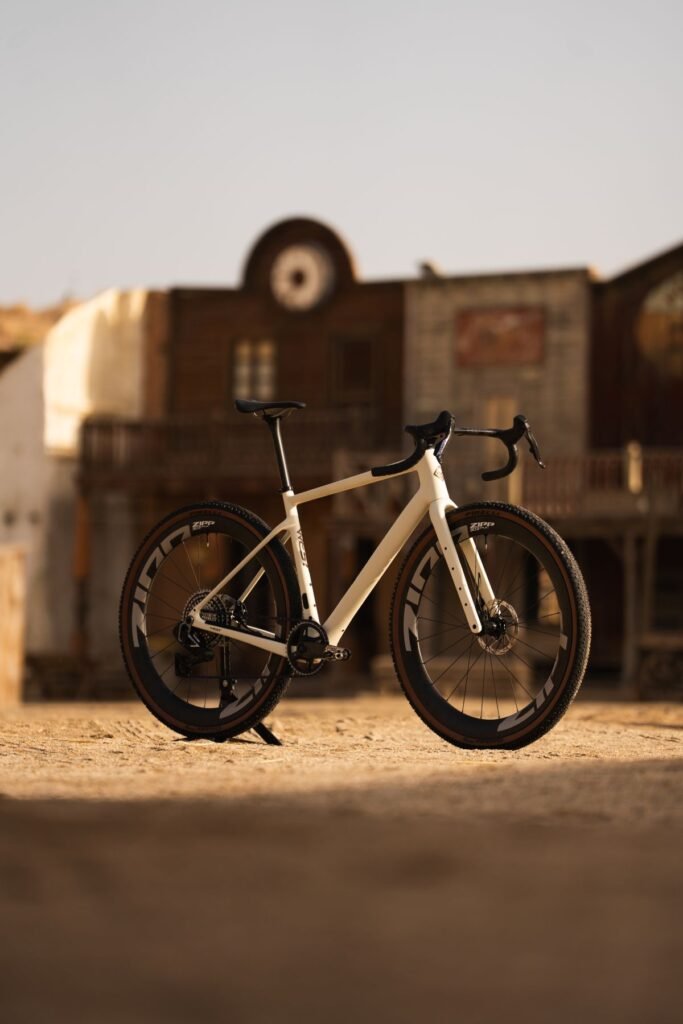 Bicicleta Megamo West 2026 en color Sand, montada con ruedas Zipp, fotografiada frente a una fachada de estilo western.